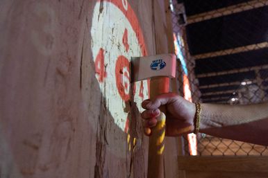 Close-up of a hand with a gold bracelet gripping an axe stuck in a splintered wooden target marked with red bullseye numbers, indoor axe-throwing lane with chain-link fence in background.