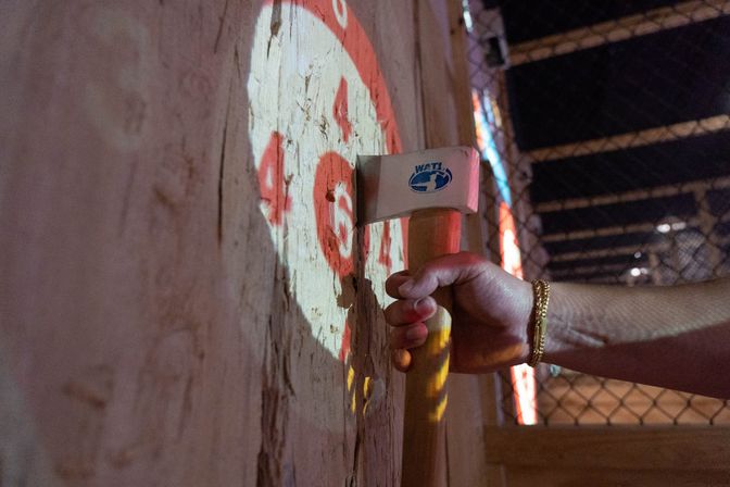 Close-up of a hand with a gold bracelet gripping an axe stuck in a splintered wooden target marked with red bullseye numbers, indoor axe-throwing lane with chain-link fence in background.