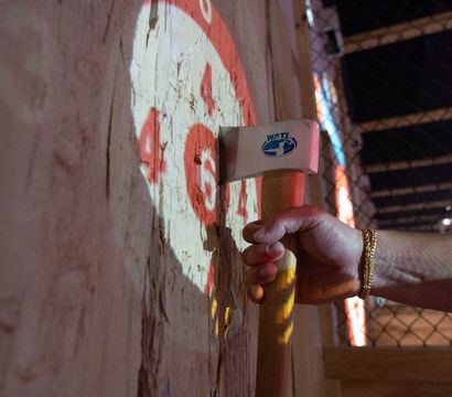 Close-up of a hand with a gold bracelet gripping an axe stuck in a splintered wooden target marked with red bullseye numbers, indoor axe-throwing lane with chain-link fence in background.