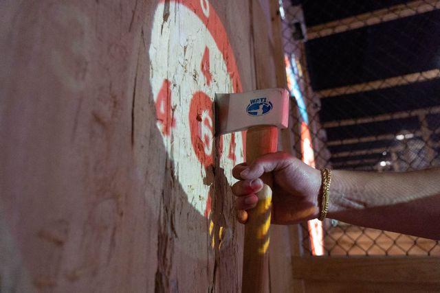 Close-up of a hand with a gold bracelet gripping an axe stuck in a splintered wooden target marked with red bullseye numbers, indoor axe-throwing lane with chain-link fence in background.