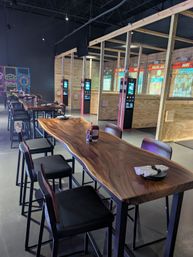 Live-edge communal wood table with high stools inside an indoor axe-throwing venue, showing fenced wooden throwing lanes with digital kiosks and colorful neon wall art in the background.