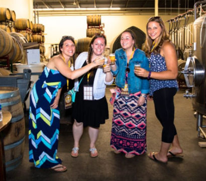 Four friends toasting with craft beers in a brewery tasting room surrounded by oak barrels and stainless steel fermentation tanks.