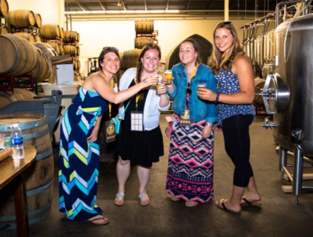 Four friends toasting with craft beers in a brewery tasting room surrounded by oak barrels and stainless steel fermentation tanks.