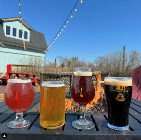 Four craft beers on a patio table by a fire pit, with string lights, picnic benches and a pale-blue building under a clear blue sky — sunny outdoor beer tasting.