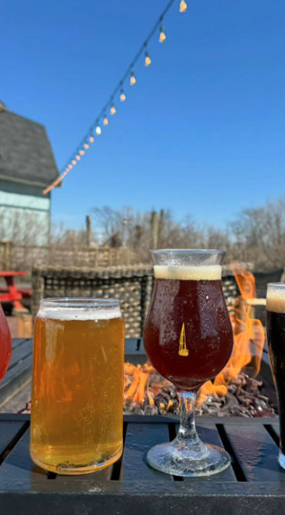Four craft beers — pink sour, golden ale, amber ale, and dark stout — lined up on a patio table by a fire pit, with string lights overhead and a blue-sky outdoor beer garden backdrop.
