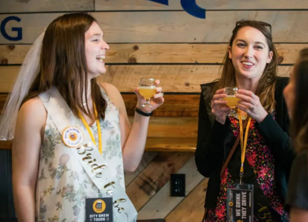 Bride-to-be and friend laughing and toasting small craft beer glasses in a rustic taproom during a bachelorette beer tasting