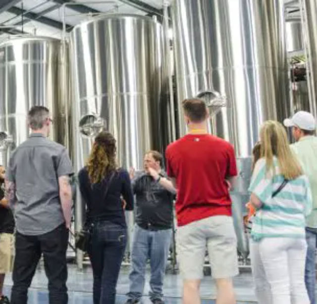 Group on a brewery tour listening to a guide in front of tall stainless-steel fermentation tanks on an indoor production floor