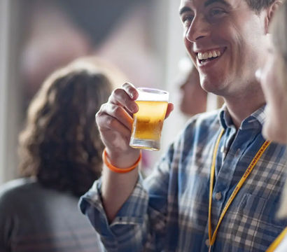 Smiling man in a plaid shirt holding a small glass of beer at an indoor craft-beer tasting or bar, lively social gathering atmosphere