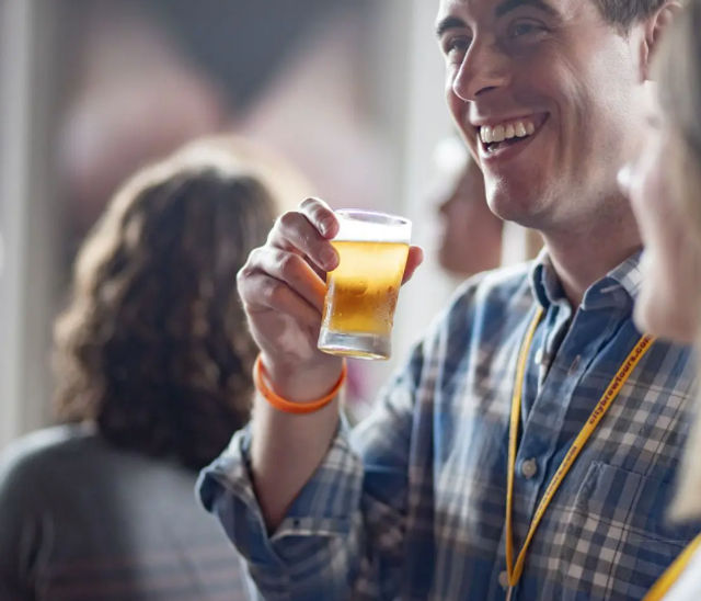 Smiling man in a plaid shirt holding a small glass of beer at an indoor craft-beer tasting or bar, lively social gathering atmosphere