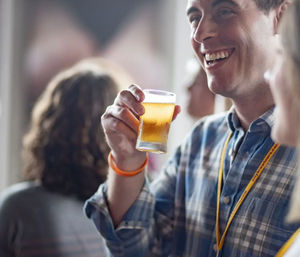 Smiling man in a plaid shirt lifting a small glass of golden craft beer at an indoor beer tasting event