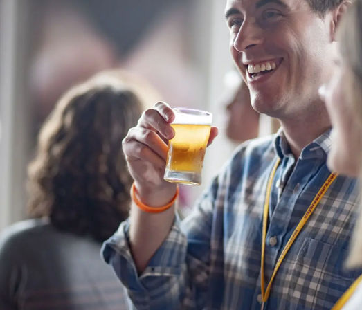 Smiling man in a plaid shirt lifting a small glass of golden craft beer at an indoor beer tasting event