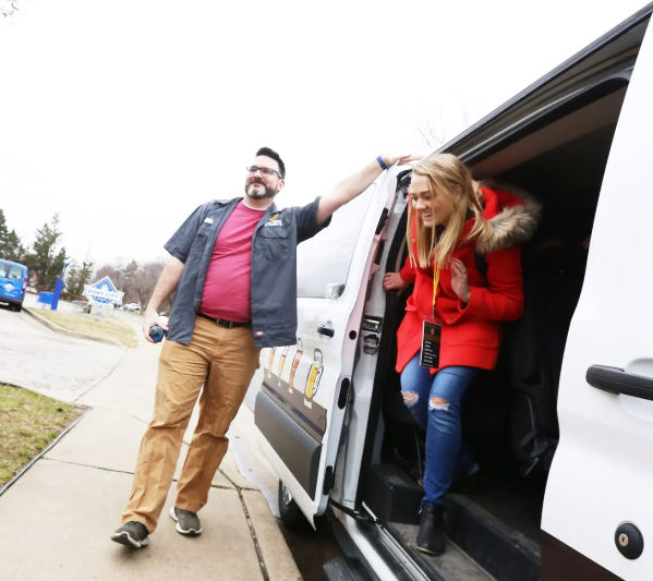 Smiling man beside an open white shuttle van on a suburban sidewalk helps a woman in a red winter coat and ripped jeans step out during a daytime neighborhood pickup.
