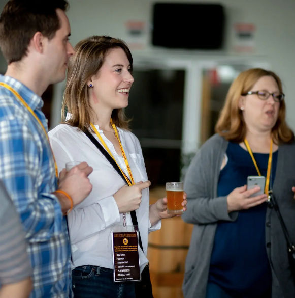 Smiling attendee holding a pint and pointing while chatting with two lanyard-wearing colleagues at an indoor networking event or craft beer tasting.