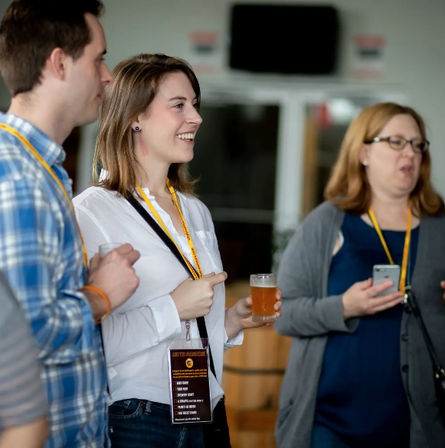Smiling attendee holding a pint and pointing while chatting with two lanyard-wearing colleagues at an indoor networking event or craft beer tasting.