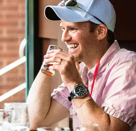 Smiling man in a light-blue cap and pink shirt sipping a sample glass of craft beer on a sunny patio