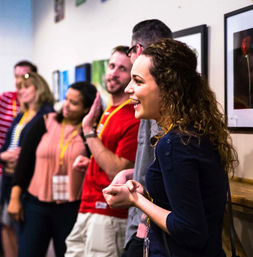 Smiling woman animatedly leading a small group of lanyard-wearing attendees during a casual networking workshop in a bright meeting room with framed photos on the wall.