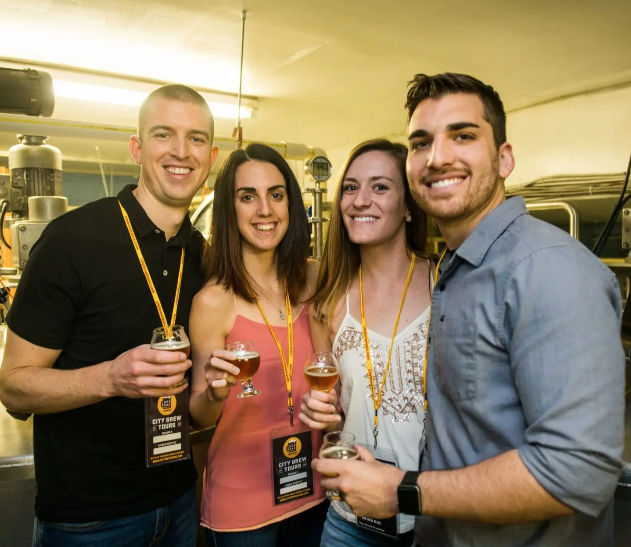 Four friends smiling and holding craft beer tasting glasses inside a microbrewery on a brewery tour, lanyards visible and stainless-steel tanks in the background.