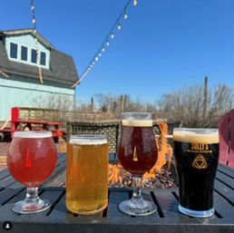 Four craft beer glasses — pink sour, pale golden ale, amber ale, and dark stout — lined up on a black patio table before a glowing fire pit, with string lights, picnic bench and a blue-sky outdoor brewery patio in the background.