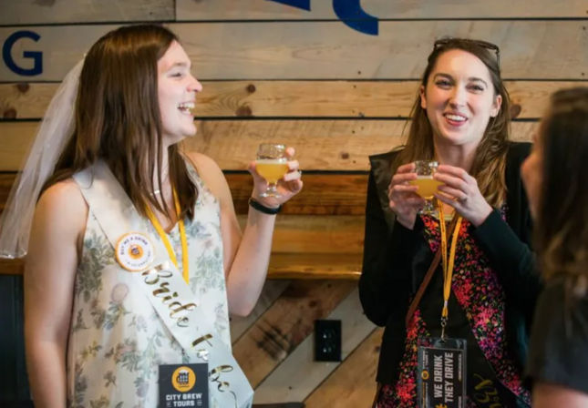 Two friends laughing and toasting small glasses of pale craft beer at a bachelorette party in a rustic brewery tasting room