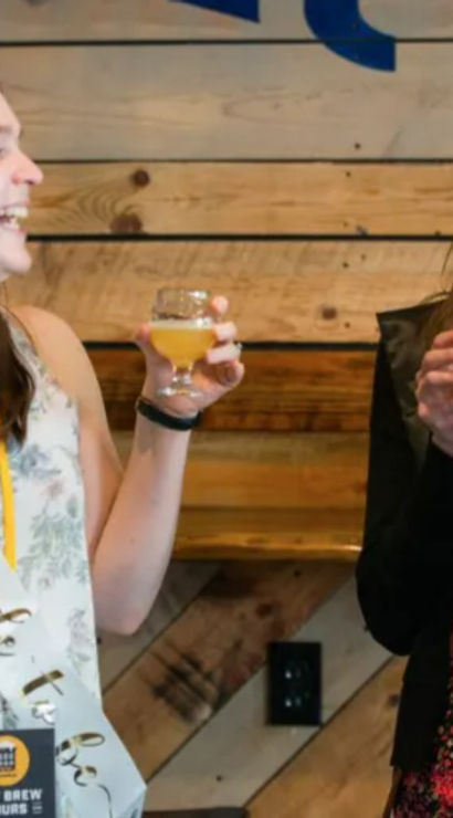 Two friends laughing and toasting small glasses of pale craft beer at a bachelorette party in a rustic brewery tasting room
