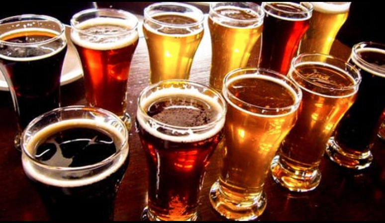 Flight of assorted craft beers in tasting glasses on a wooden bar table, showcasing dark stouts, amber ales and pale golden lagers.