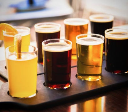 Eight-sample craft beer flight on a wooden tasting paddle — pale, amber and dark pours with a citrus garnish in a taproom