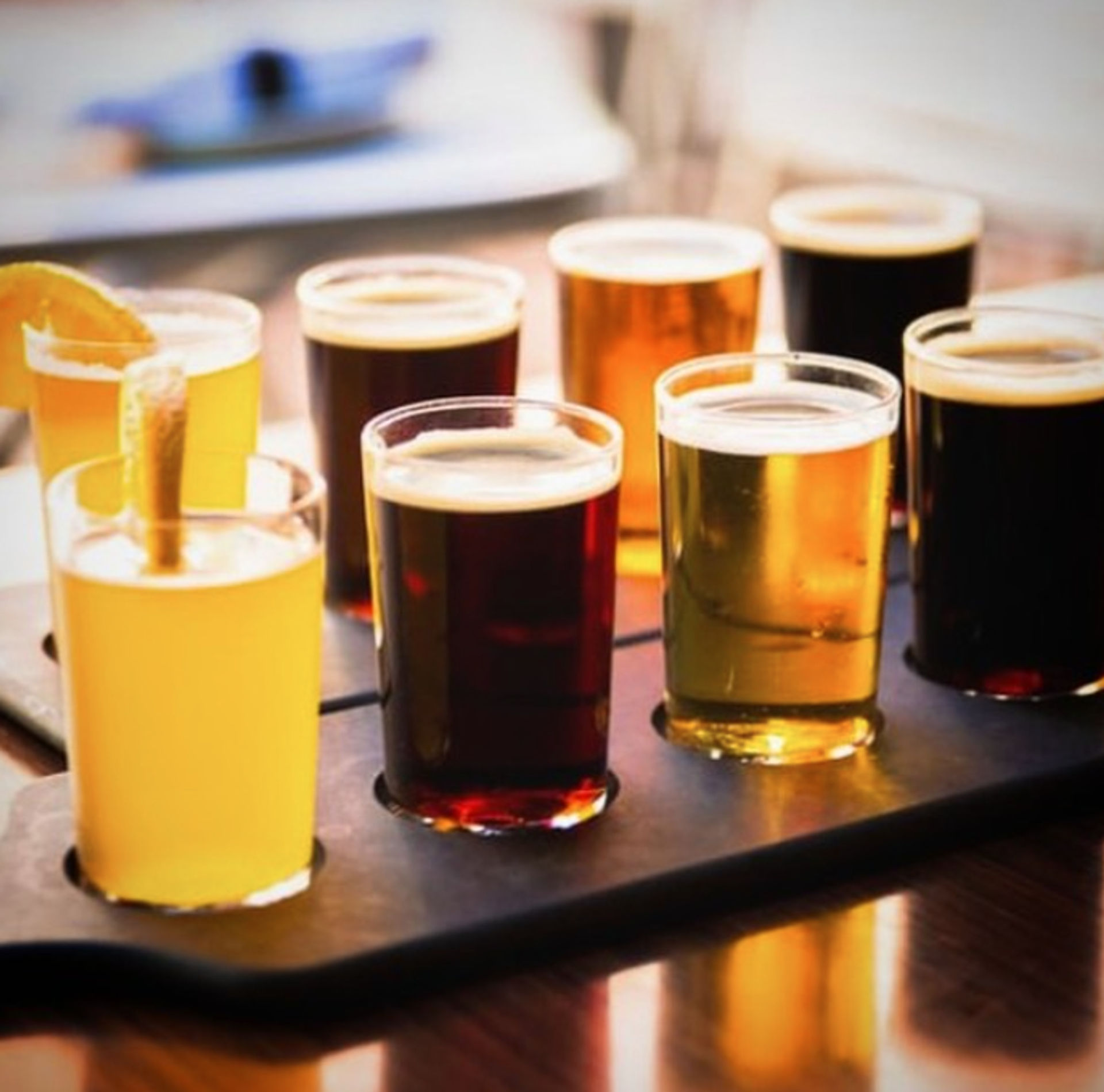 Eight-sample craft beer flight on a wooden tasting paddle — pale, amber and dark pours with a citrus garnish in a taproom