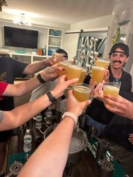 Indoor house party toast — friends clinking short citrus cocktails over an ice bucket in a living-room bar area with silver foil balloons in the background.