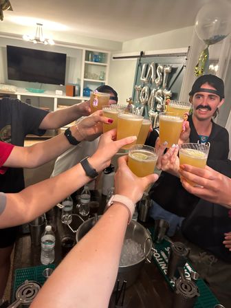 Indoor house party toast — friends clinking short citrus cocktails over an ice bucket in a living-room bar area with silver foil balloons in the background.