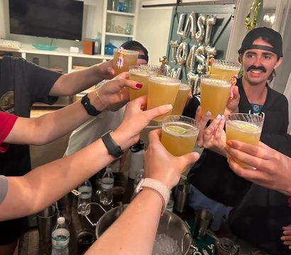 Indoor house party toast — friends clinking short citrus cocktails over an ice bucket in a living-room bar area with silver foil balloons in the background.