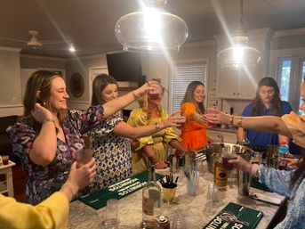 Group of women toasting with cocktails around a marble kitchen island under pendant lights at a lively home cocktail party, with bartending tools and bottles on the counter.