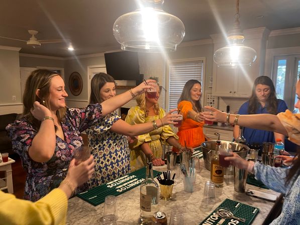 Group of women toasting with cocktails around a marble kitchen island under pendant lights at a lively home cocktail party, with bartending tools and bottles on the counter.