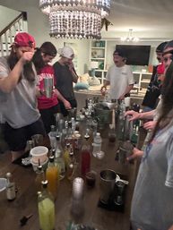 Friends at a living room house party gathered around a table turned cocktail station with shakers, bottles, mixers and plastic cups beneath a beaded chandelier.