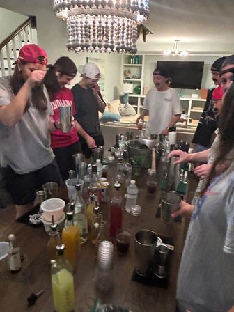 Friends at a living room house party gathered around a table turned cocktail station with shakers, bottles, mixers and plastic cups beneath a beaded chandelier.