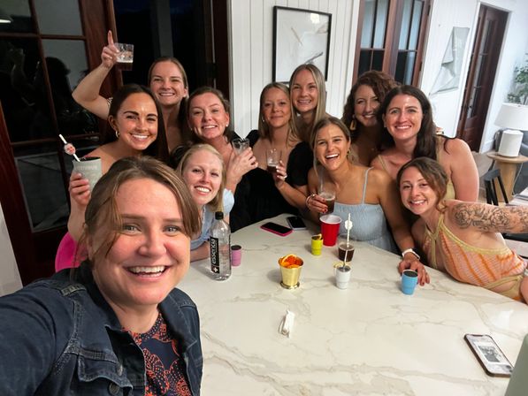 Cheerful group of friends taking a selfie around a marble kitchen island in a bright modern home, smiling and holding drinks and colorful cups at an indoor party.