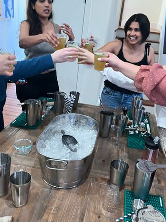 Group of friends toasting iced cocktails over a wooden table with a stainless ice bucket and bartending tools in a home kitchen.