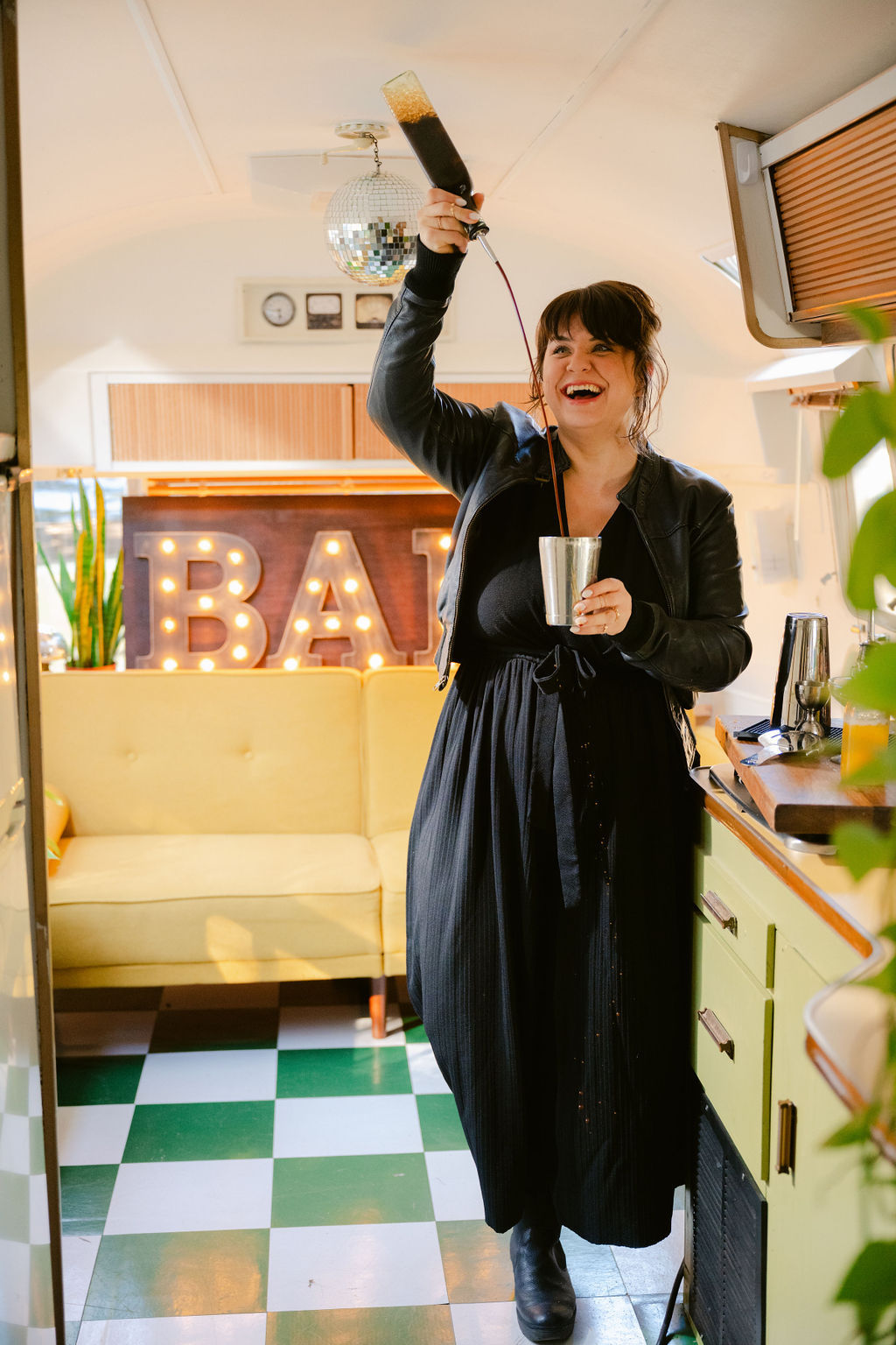 Smiling bartender pouring a dark cocktail from a bottle into a metal shaker inside a retro camper mobile bar with illuminated "BAR" marquee, disco ball, yellow sofa and green-and-white checkerboard floor.
