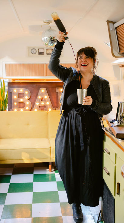 Smiling bartender pouring a dark cocktail from a bottle into a metal shaker inside a retro camper mobile bar with illuminated "BAR" marquee, disco ball, yellow sofa and green-and-white checkerboard floor.
