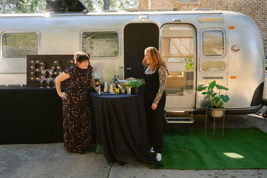 Two women chatting at an outdoor patio cocktail station in front of a silver vintage camper converted into a mobile bar, with a marquee 'BAR' sign, high-top table holding mixers and glassware, potted plants and green turf.