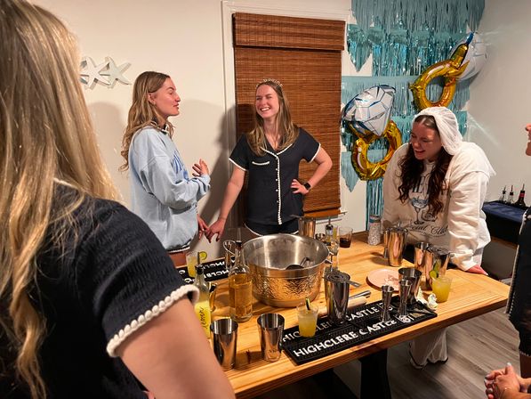 Friends laughing around a wooden table turned cocktail bar at a cozy bachelorette party, bride-to-be wearing a tiara and ring-shaped balloons in the background.