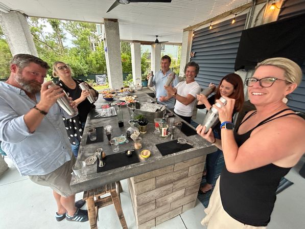 Smiling group shaking cocktail shakers around a covered outdoor patio bar with drinks, snacks and string lights.