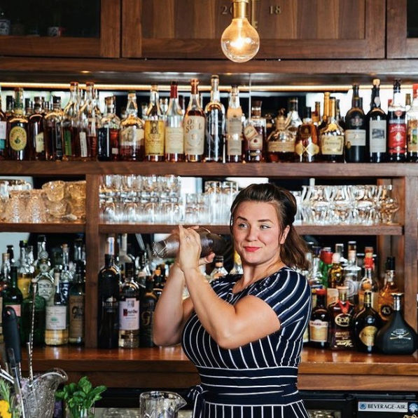 Smiling bartender shaking a cocktail shaker at a wooden back bar lined with liquor bottles and glassware under a warm pendant light.