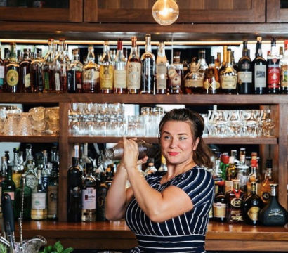 Smiling bartender shaking a cocktail shaker at a wooden back bar lined with liquor bottles and glassware under a warm pendant light.