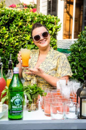 Smiling person in sunglasses at a backyard garden cocktail station holding a layered orange-yellow cocktail garnished with mint, surrounded by bottles, fresh herbs and ice-filled glasses.