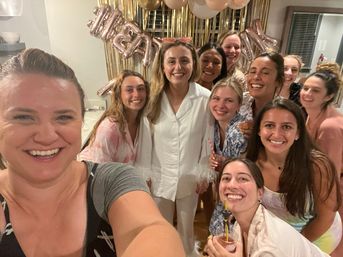 Group selfie of smiling women at an indoor pajama-style celebration with gold fringe backdrop, pink and gold balloons and friends holding drinks — fun girls' night party scene