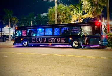 Neon-lit party bus parked on a palm-lined street at night, black exterior glowing with blue and pink lights and palm graphics, evoking a lively coastal nightlife vibe.
