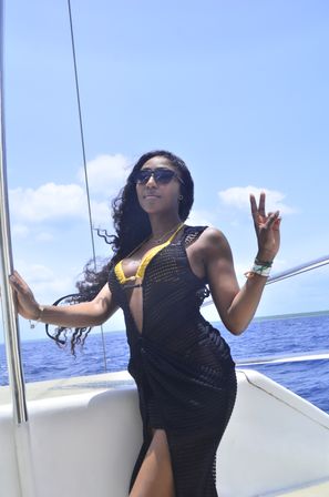 Sun-soaked woman in a yellow bikini top and black mesh cover-up wearing sunglasses, flashing a peace sign aboard a boat with deep blue ocean and clear sky — tropical sailing vibe.