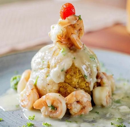 Mouthwatering Puerto Rican-style mofongo: a golden plantain mound draped in creamy garlic sauce, ringed with sautéed garlic shrimp and crowned with a bright cherry tomato on a blue plate.