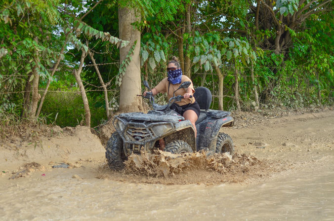 ATV rider wearing a bandana and sunglasses splashing through a muddy puddle on a tropical forest dirt trail