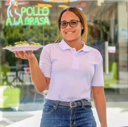 Smiling server in a white polo and jeans holding a plate of grilled chicken and salad in front of a restaurant window with a neon "Pollo a la Brasa" sign.